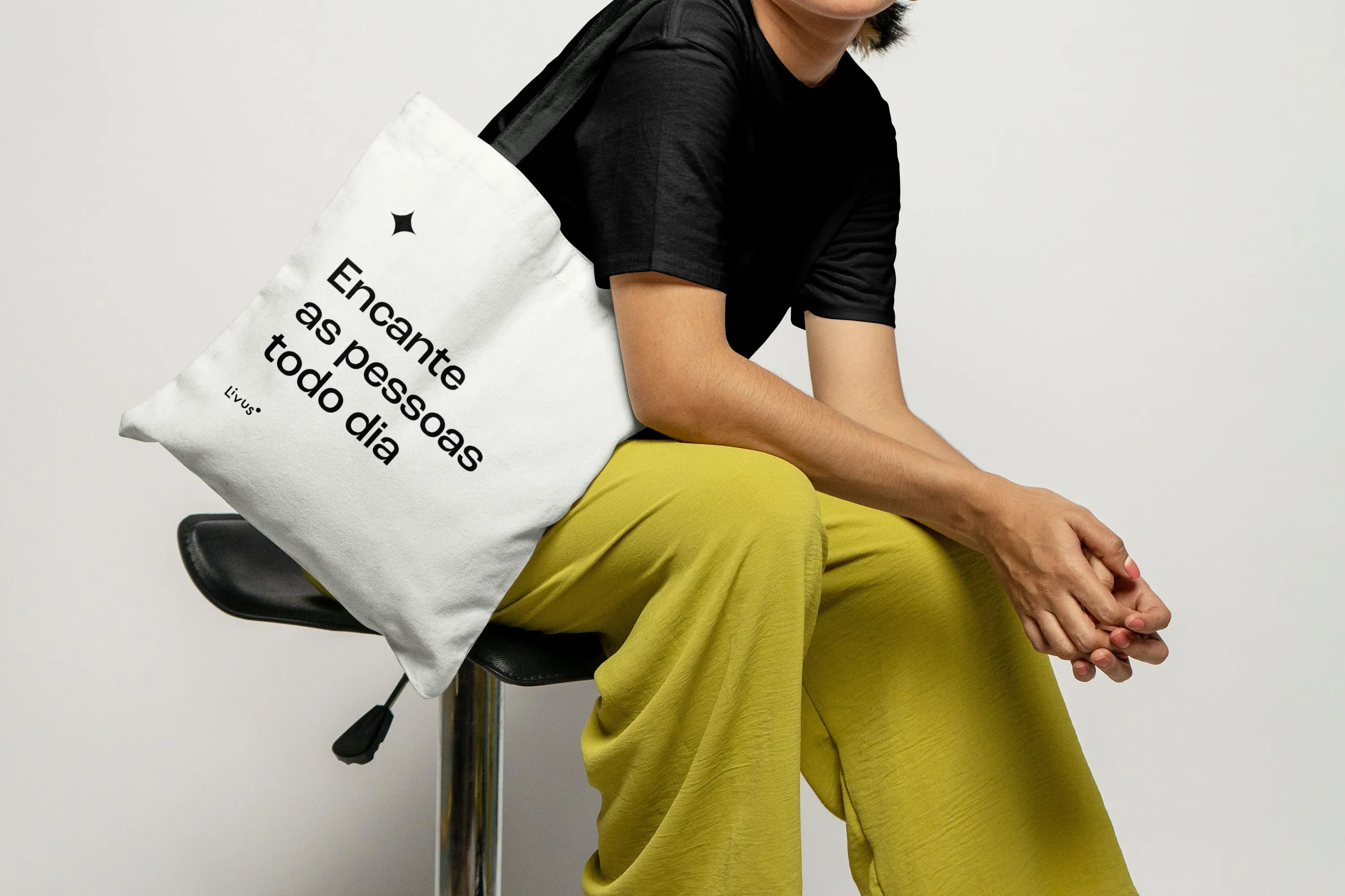 A white Livus tote bag hanging from the shoulder of a seated person, printed with a black star and the line "Encante as pessoas todo dia".