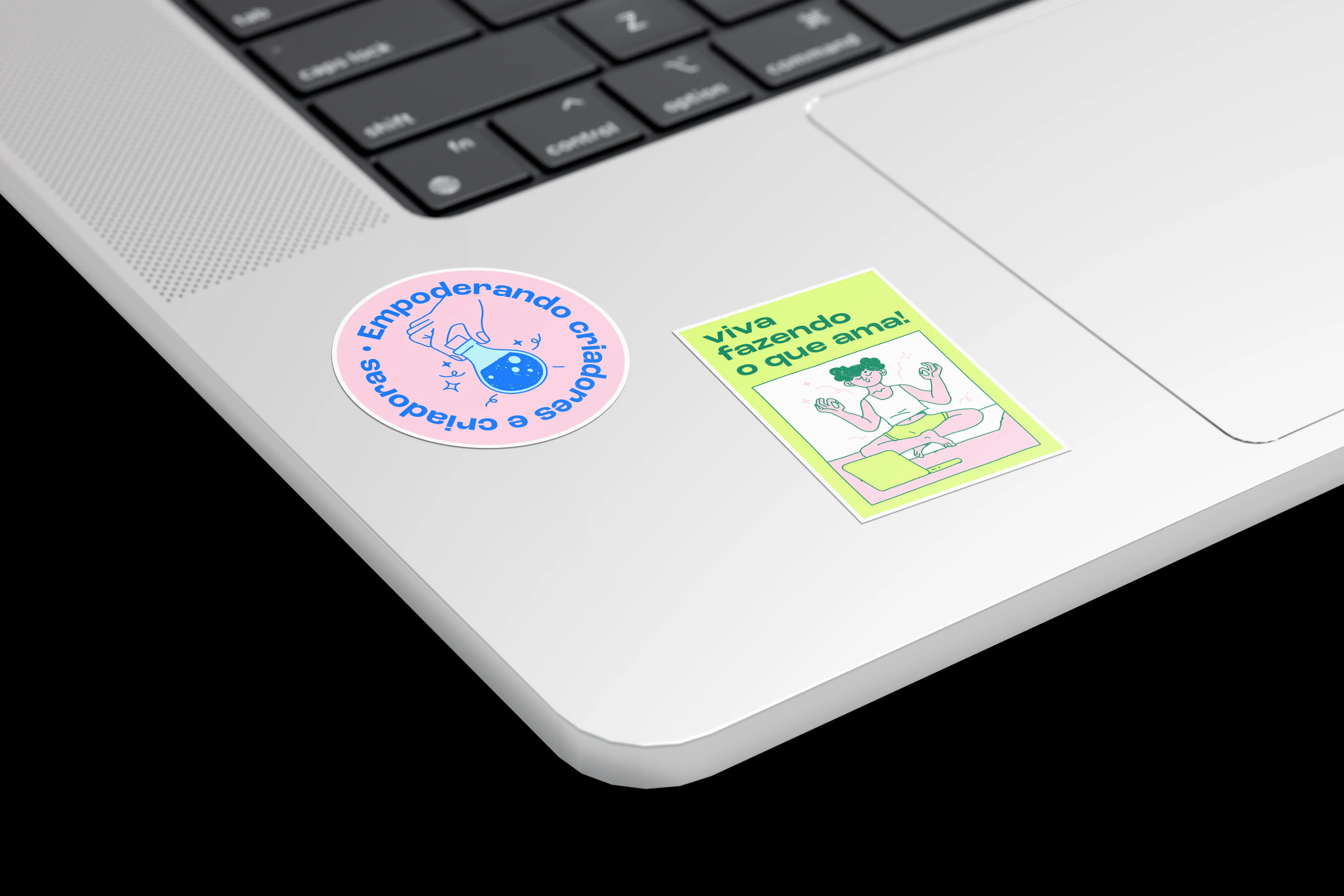 Two stickers on a silver laptop beside the trackpad: a pink circle reading "Empoderando criadores e criadoras" and a lime card reading "Viva fazendo o que ama!" with a meditating figure.