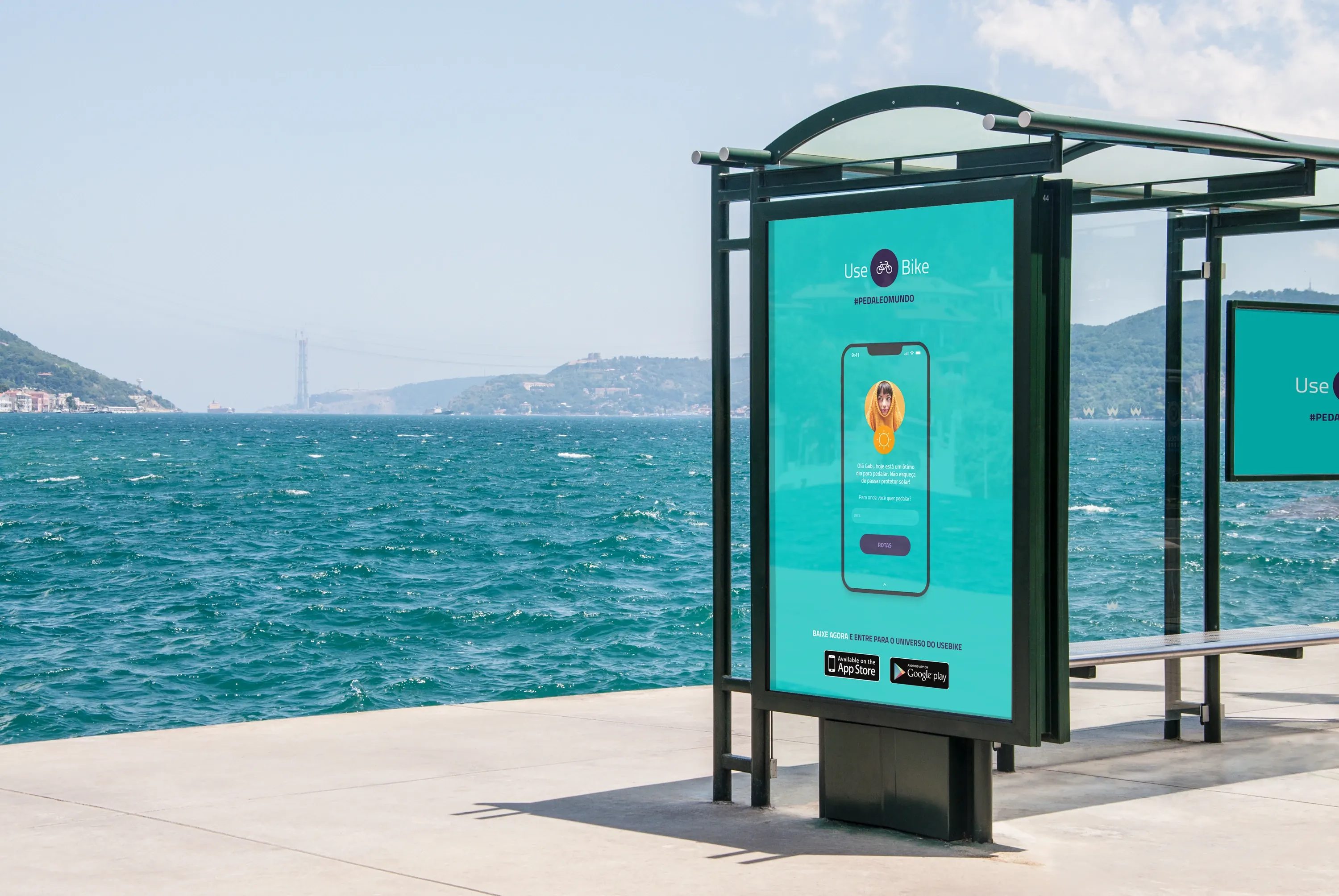 A teal UseBike ad in a seaside bus shelter, showing the app's greeting screen on a phone above "Baixe agora" and App Store and Google Play badges.