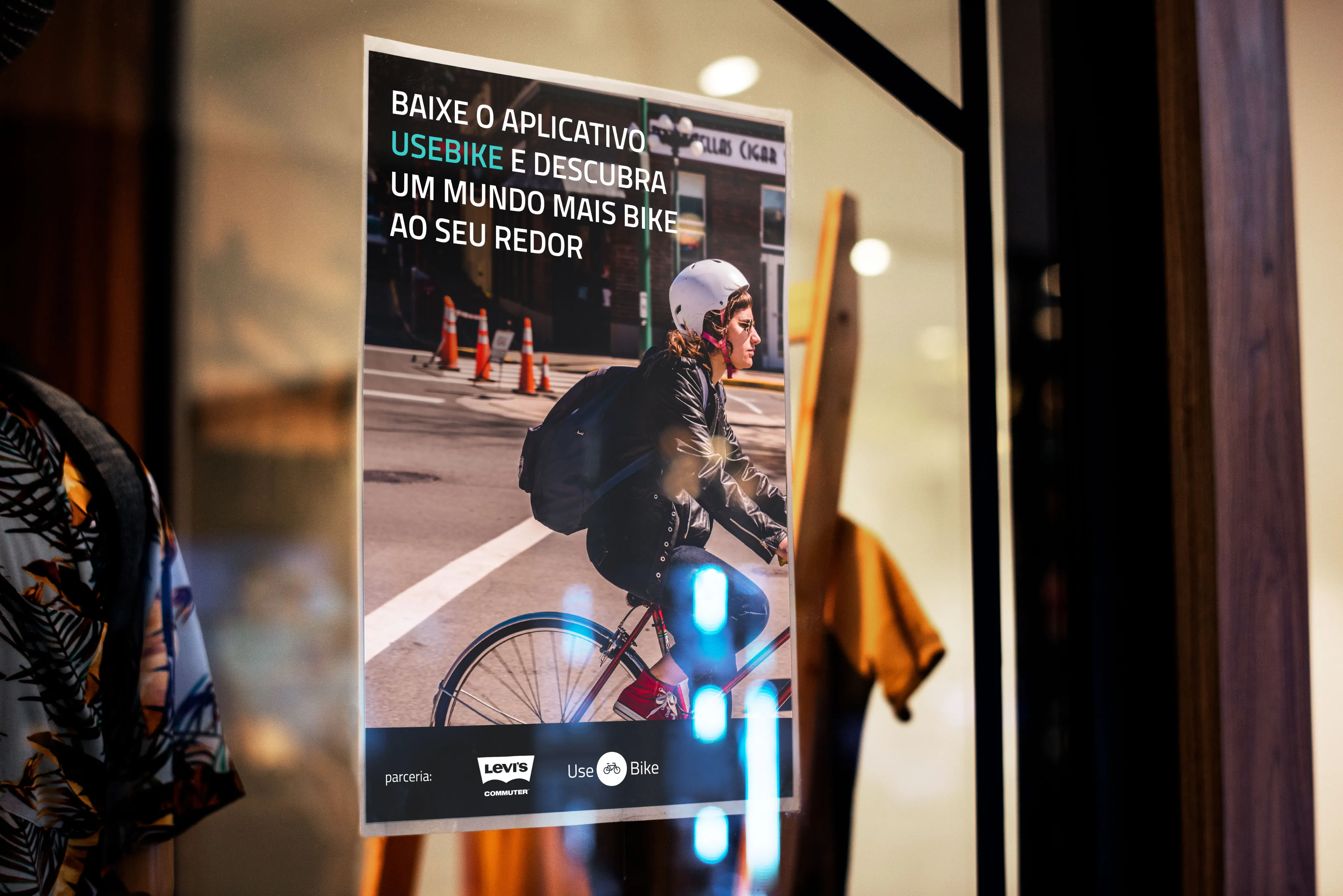 A poster in a shop window, a woman in a helmet cycling across a street under the headline "Baixe o aplicativo UseBike", with Levi's Commuter and UseBike logos along the bottom.