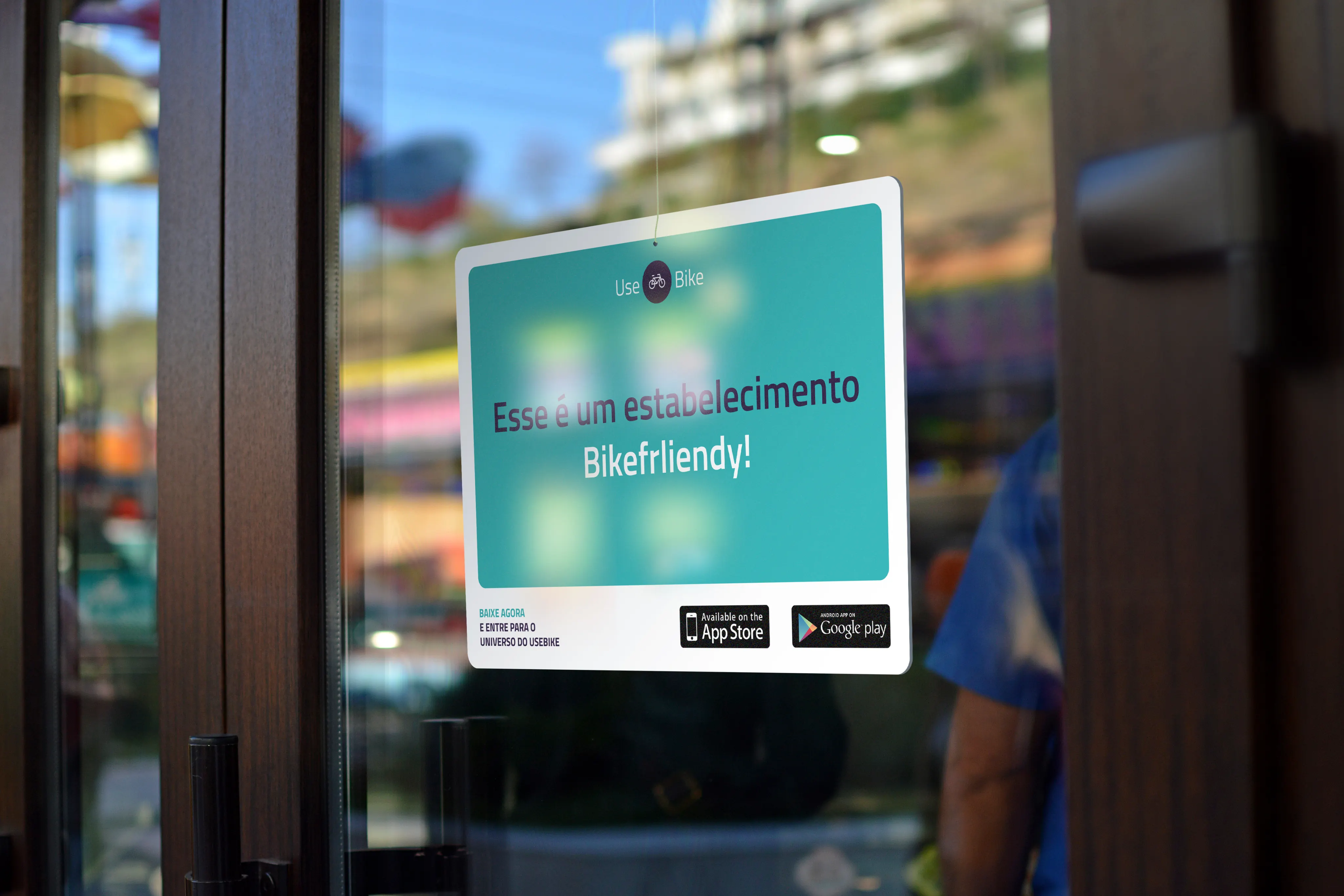 A teal and white UseBike sign hanging on a glass shop door, telling passers-by the business is bike-friendly, with App Store and Google Play badges.