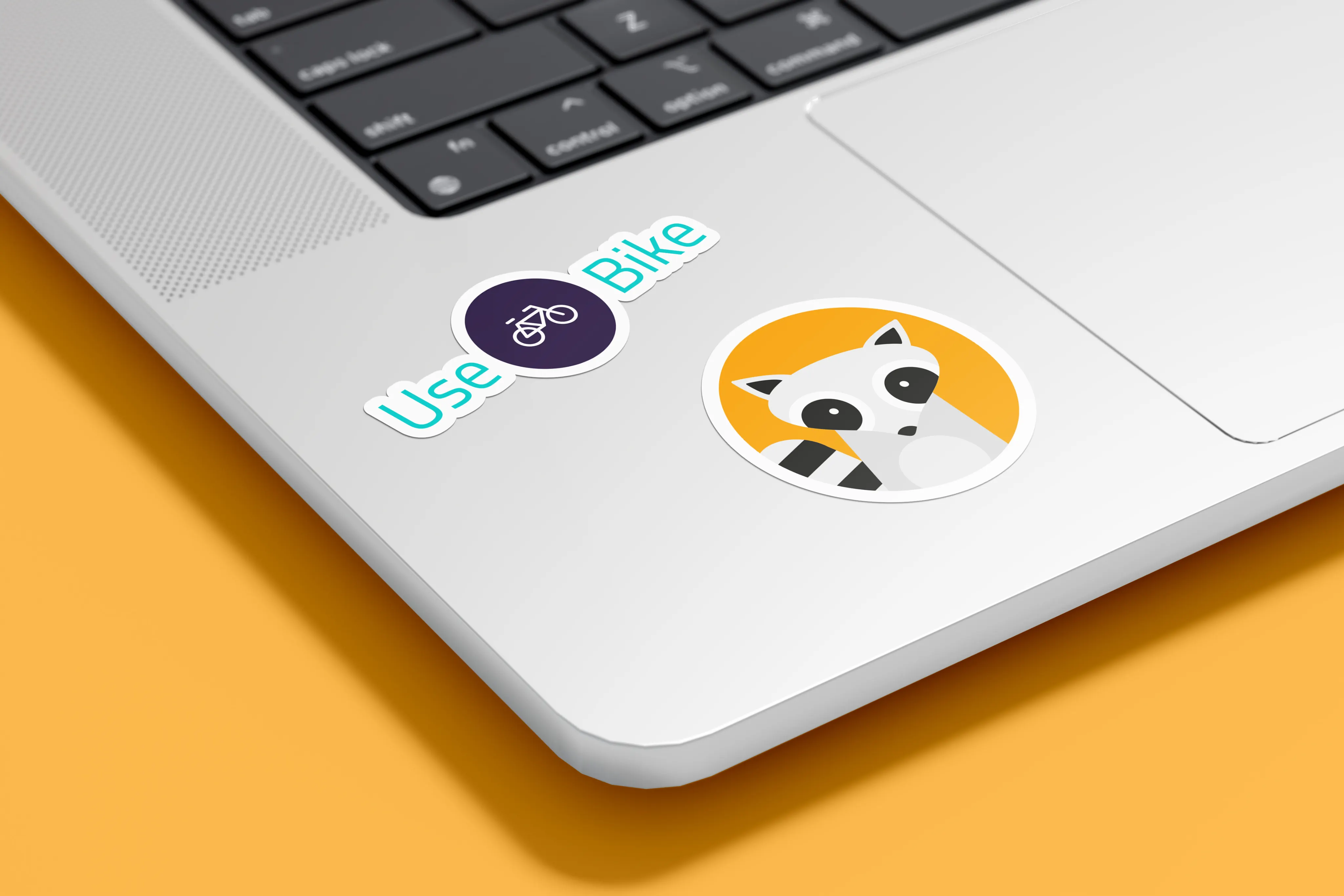 Two stickers on a silver laptop palm rest against an orange background: the UseBike wordmark and the round raccoon mascot.